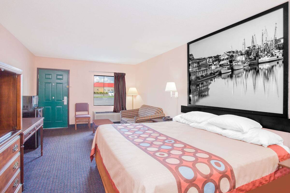A hotel room with a bed, a desk, a chair, a window with curtains, a lamp, and a large framed picture on the wall.