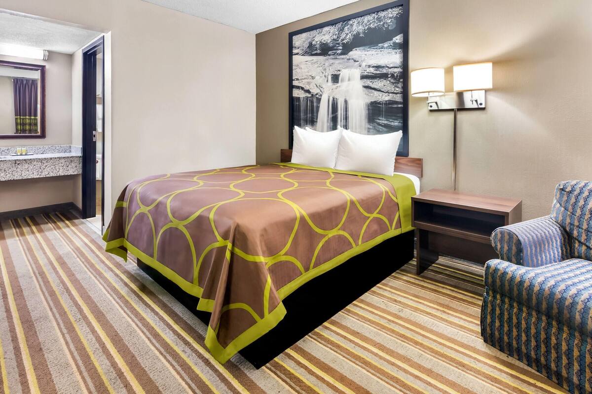 A hotel room with a bed, a chair, a nightstand, a lamp, and a painting on the wall.