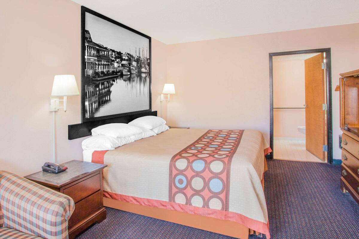 A hotel room with a bed, two bedside lamps, a nightstand, a chair, a wooden dresser, and a door leading to another room.