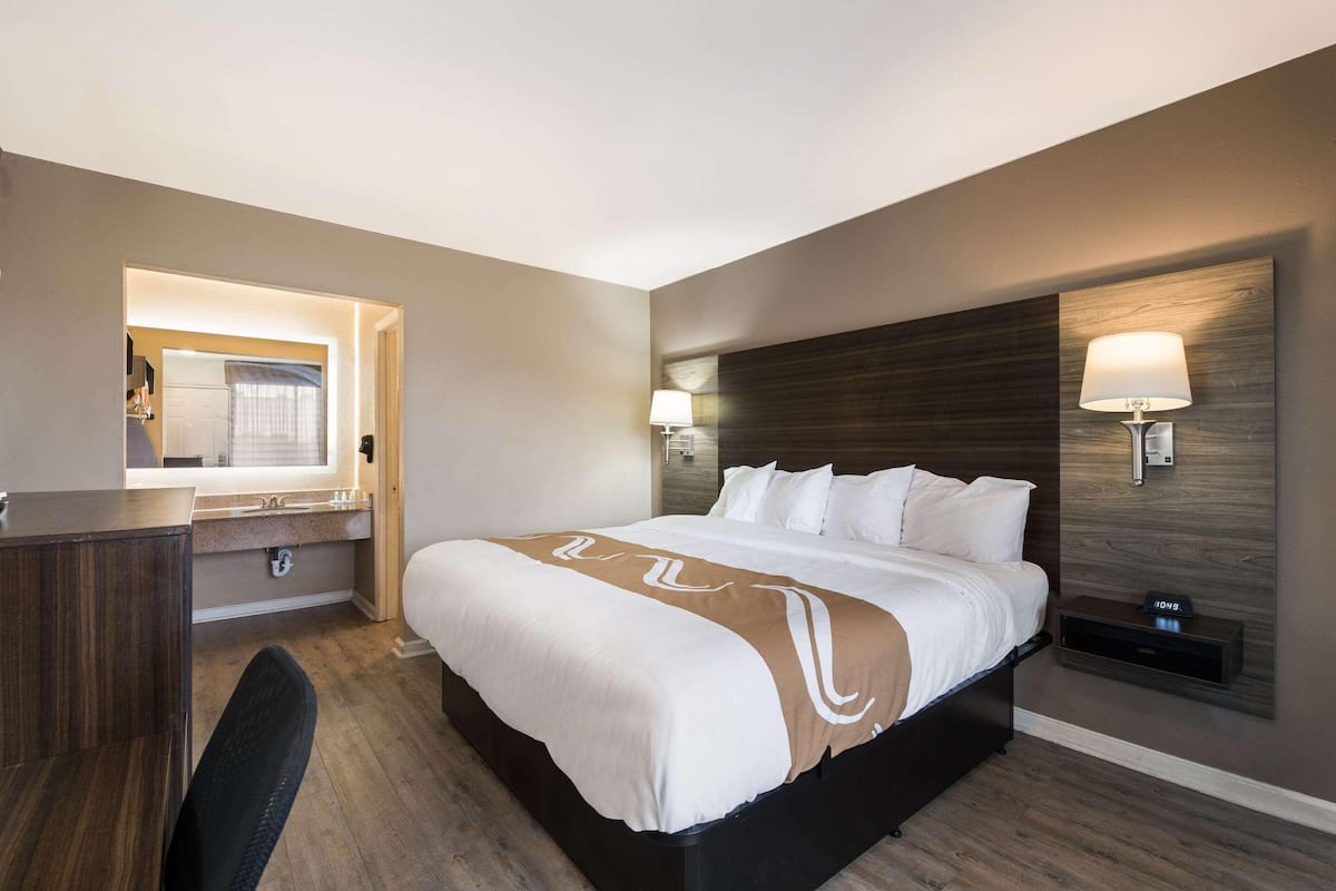 A hotel room with a bed, a desk, a chair, a mirror, and two wall-mounted lamps.