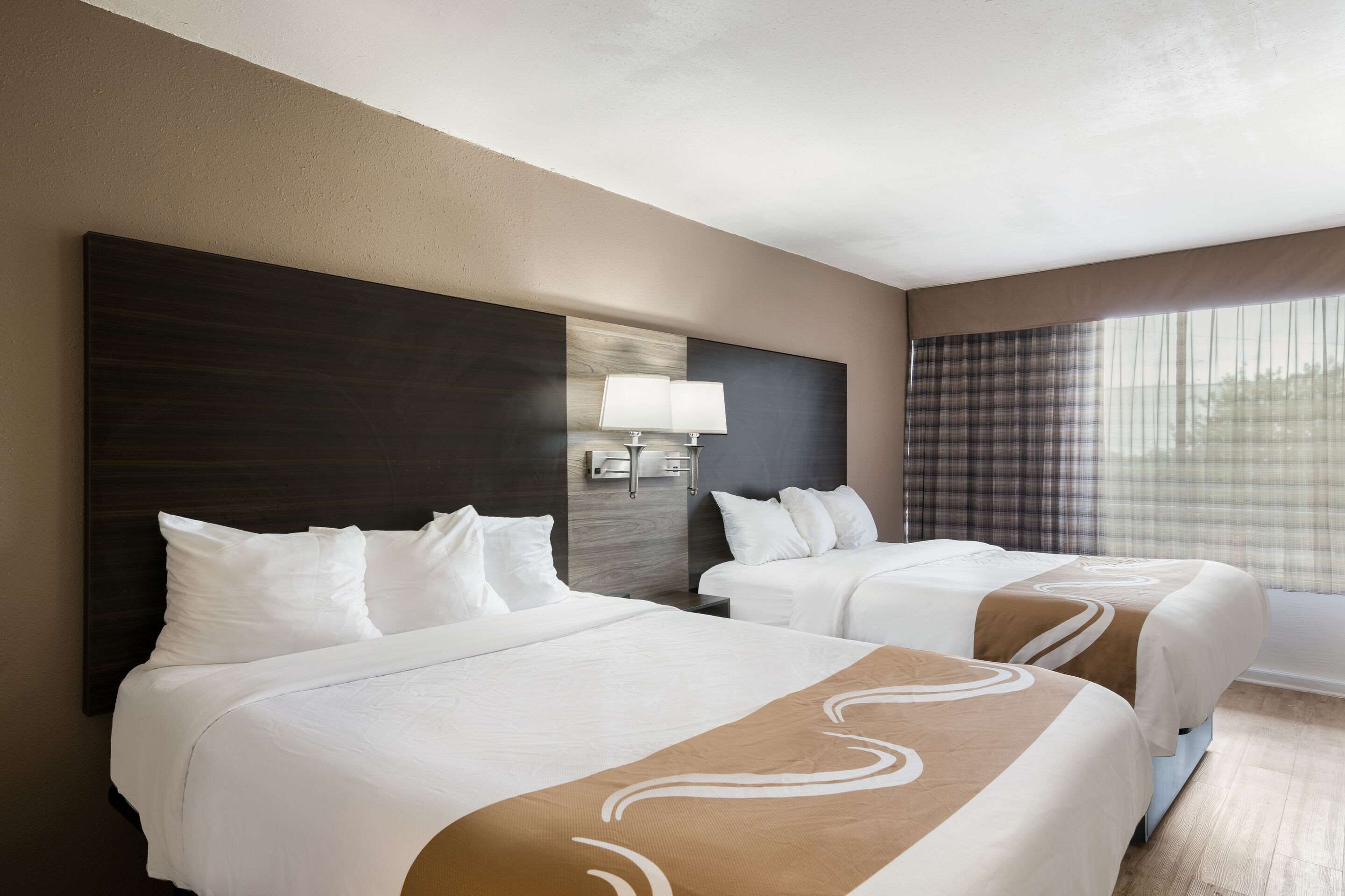 A hotel room with two beds, a large dark headboard, a wall-mounted lamp, and a window with curtains.