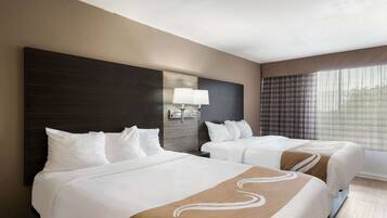 A hotel room with two beds, a large dark headboard, a wall-mounted lamp, and a window with curtains.