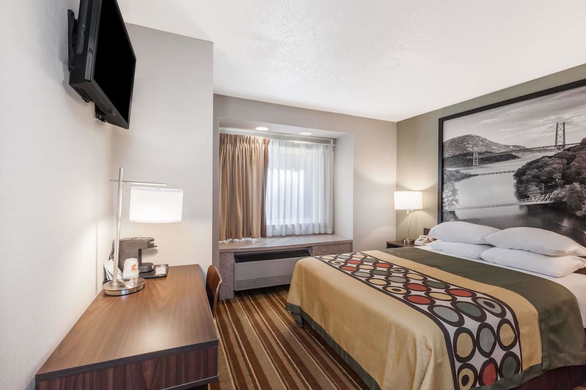 A hotel room with a bed, a desk, a chair, a TV mounted on the wall, a window with curtains, and a painting on the wall.
