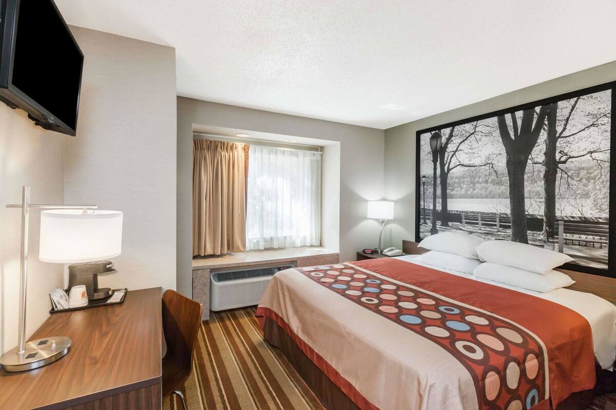 A hotel room with a bed, a desk, a chair, a TV, a window with curtains, a lamp, and a painting on the wall.