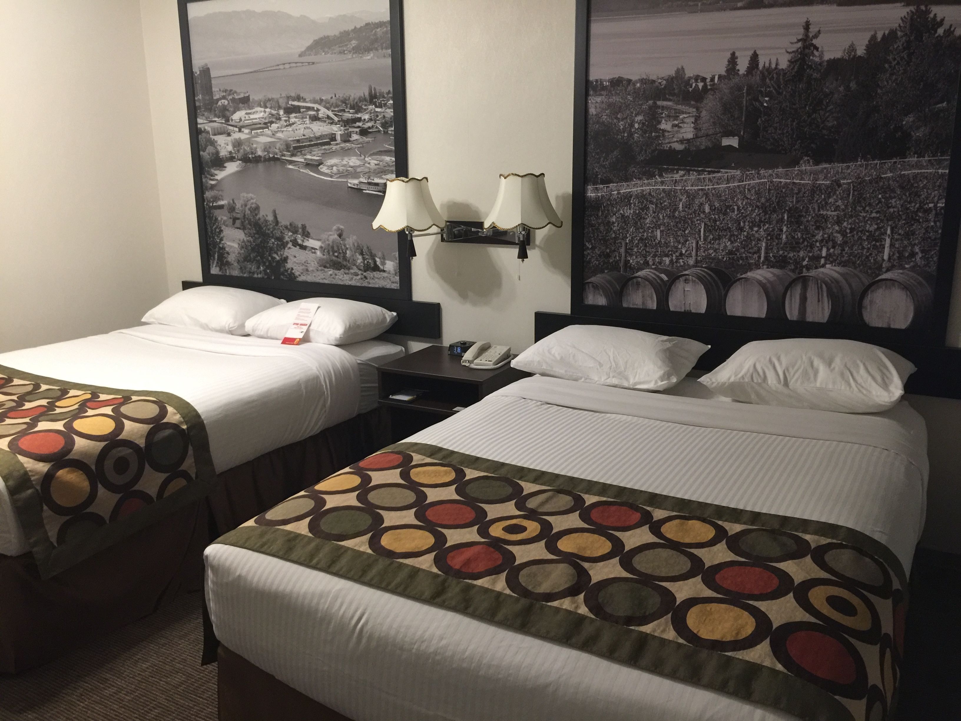 A hotel room with two beds, each with a different patterned bedspread. There are two wall-mounted lamps and a nightstand with a phone. The room features large black-and-white landscape pictures on the walls.