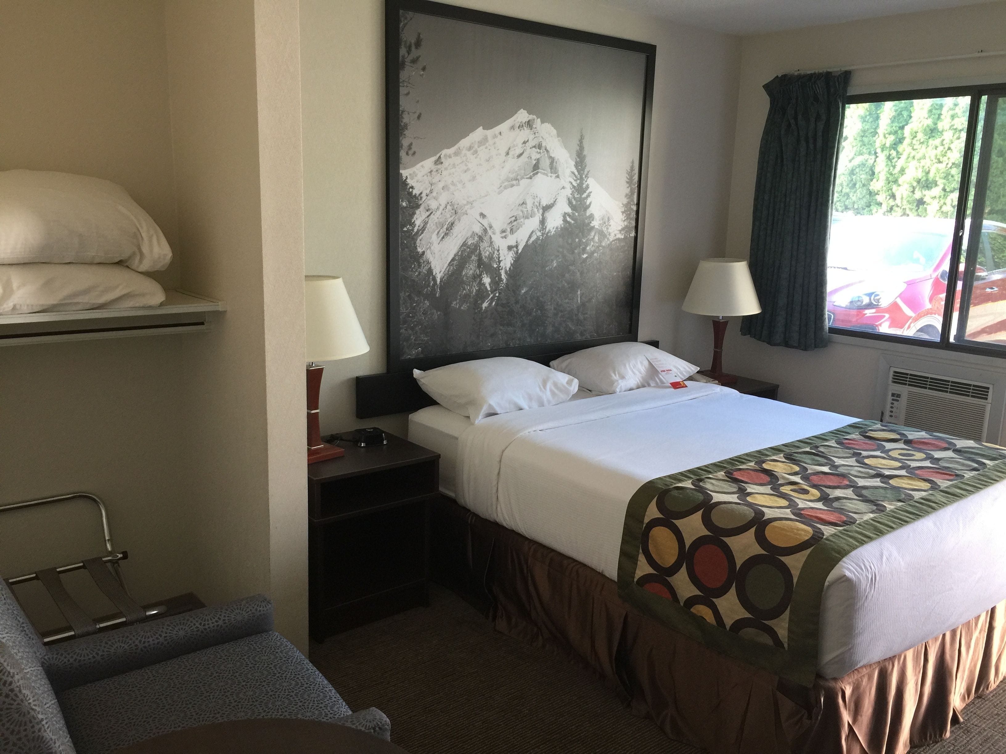 Habitación de hotel con una cama, dos mesitas de noche, una silla, un cuadro grande con una imagen de montaña y una ventana con cortinas.