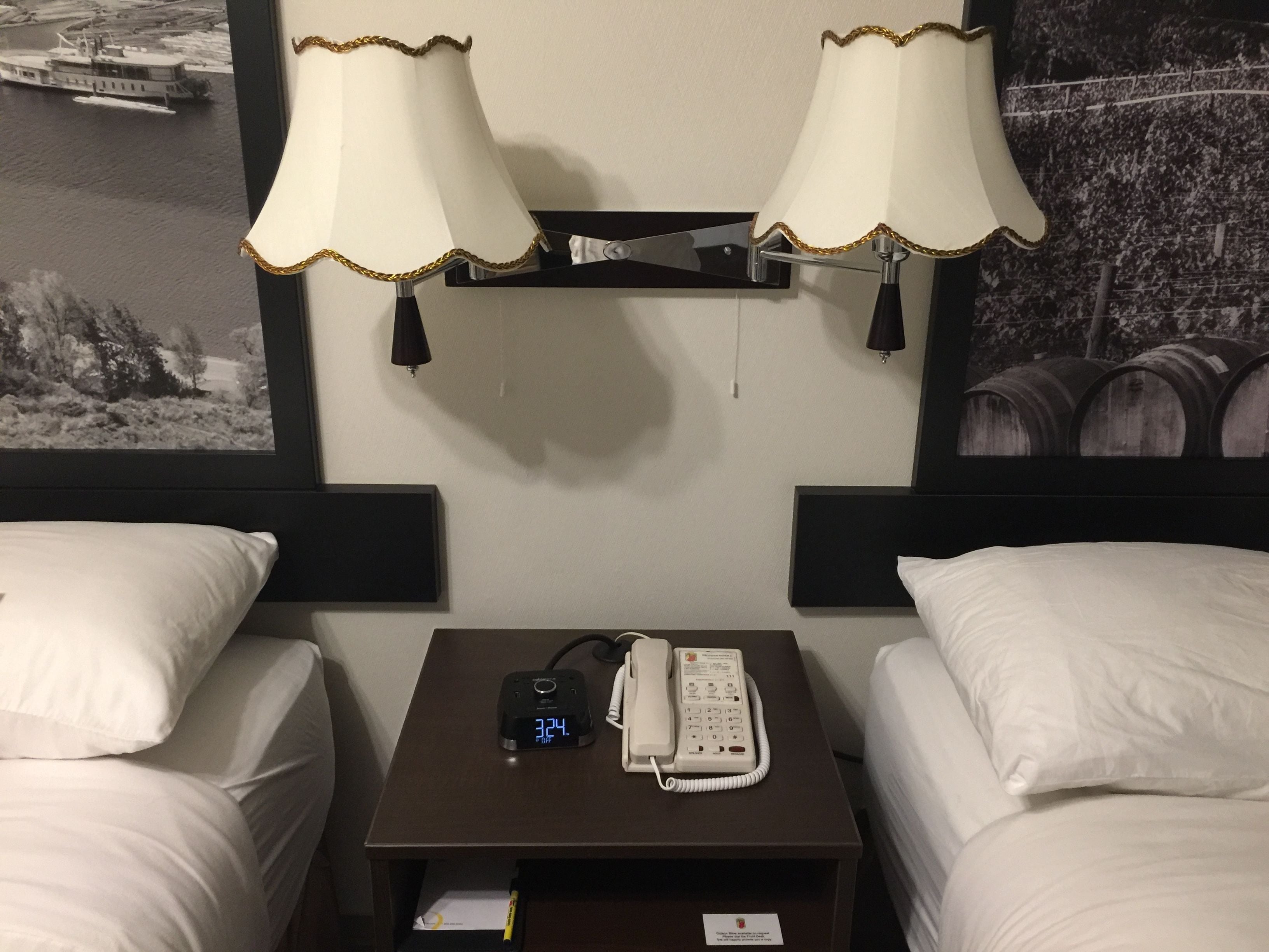 A hotel room with two beds, a nightstand with a digital clock and a telephone, and two wall-mounted lamps.
