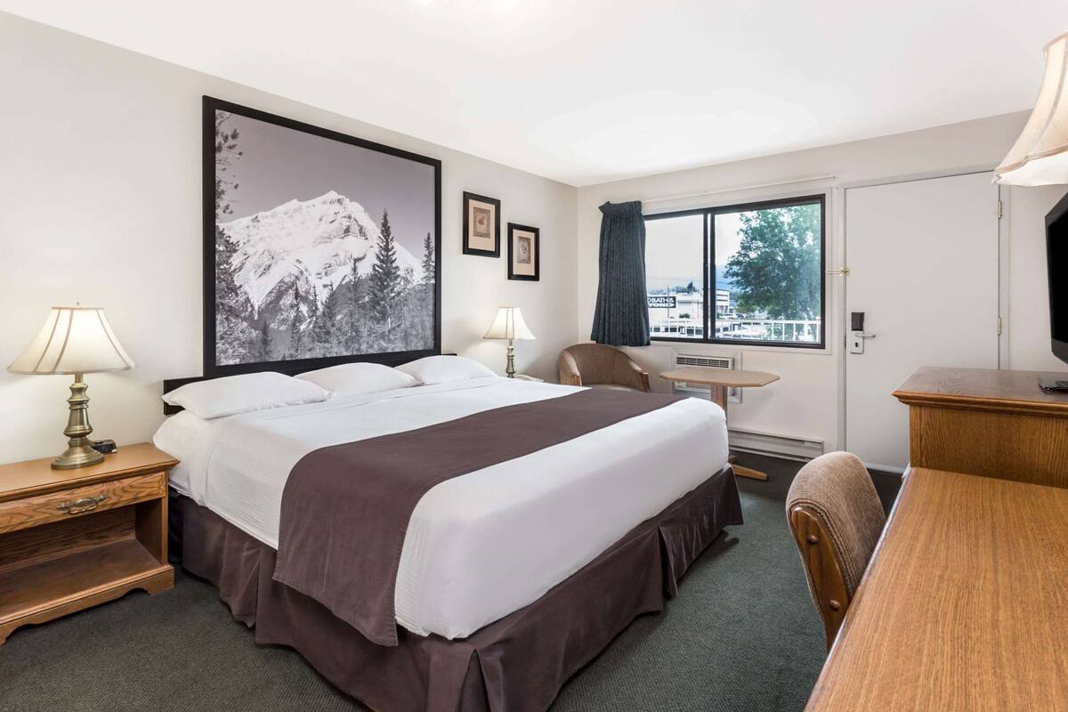 A hotel room with a bed, bedside tables, a desk, a chair, a window with a view, and a large framed picture on the wall.