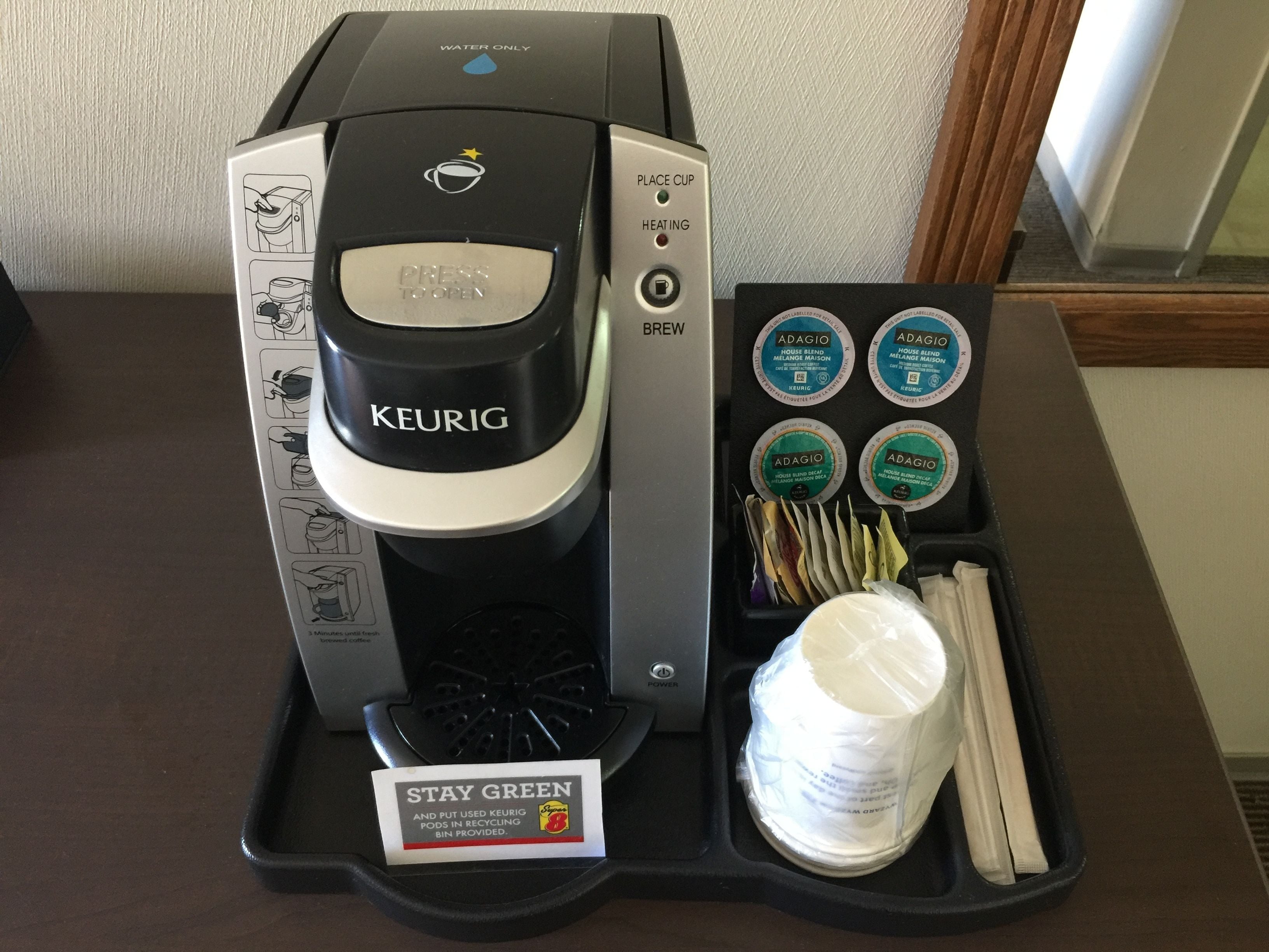 Una cafetera Keurig con una bandeja que contiene tazas reutilizables, tapas y un contenedor de reciclaje 'Stay Green'.