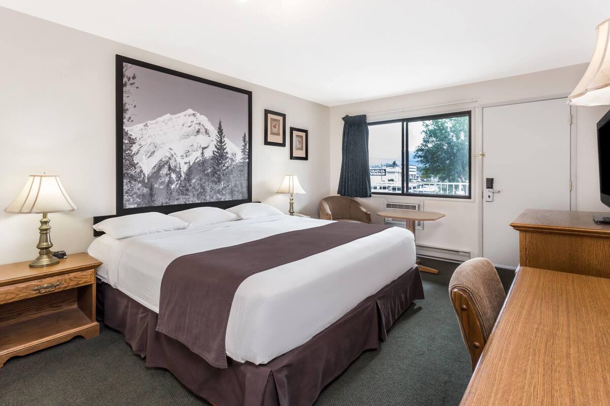 A hotel room with a bed, bedside tables, a desk, a chair, a window with a view, and a large framed picture on the wall.