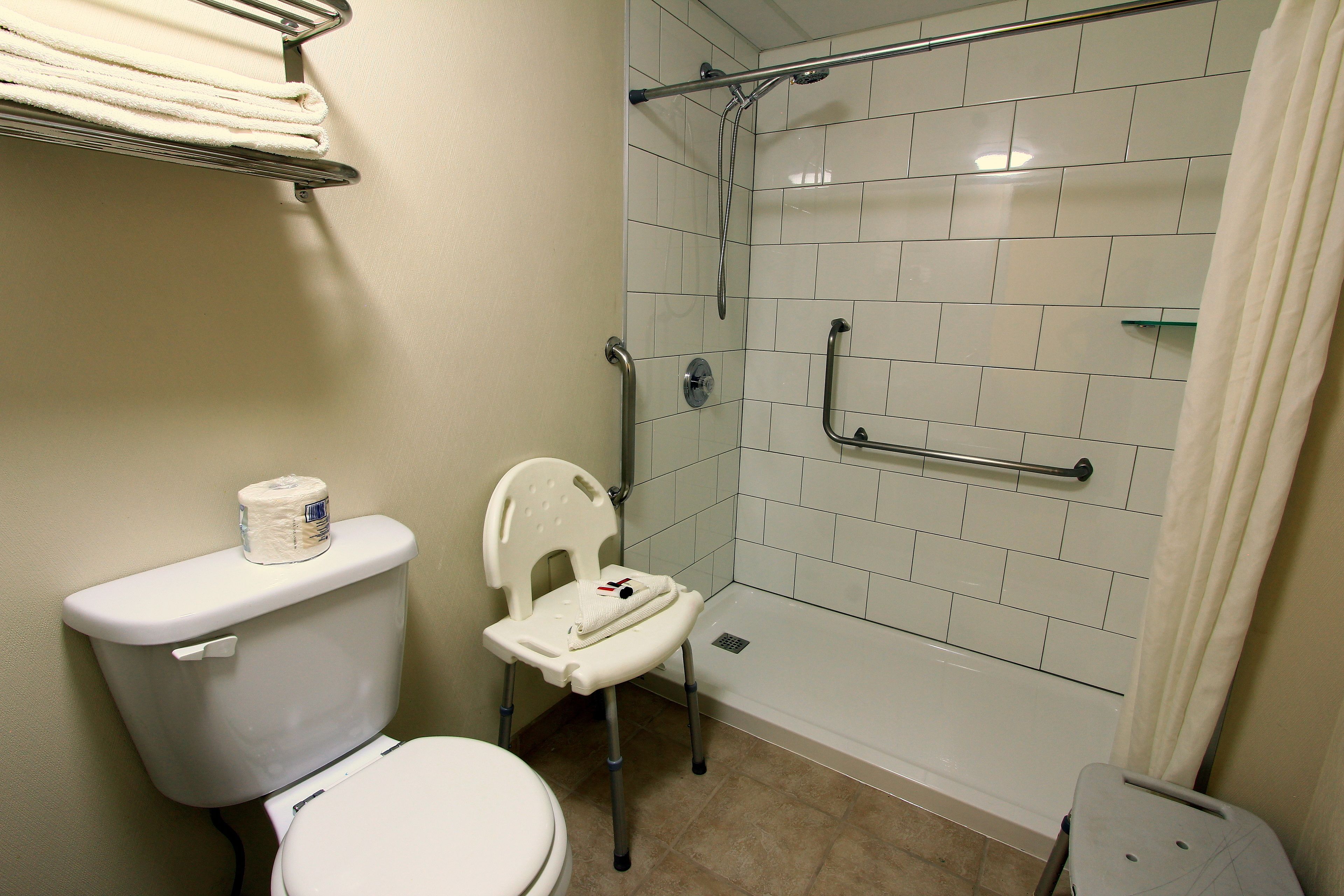 A bathroom with a toilet, a shower with grab bars, and a chair.