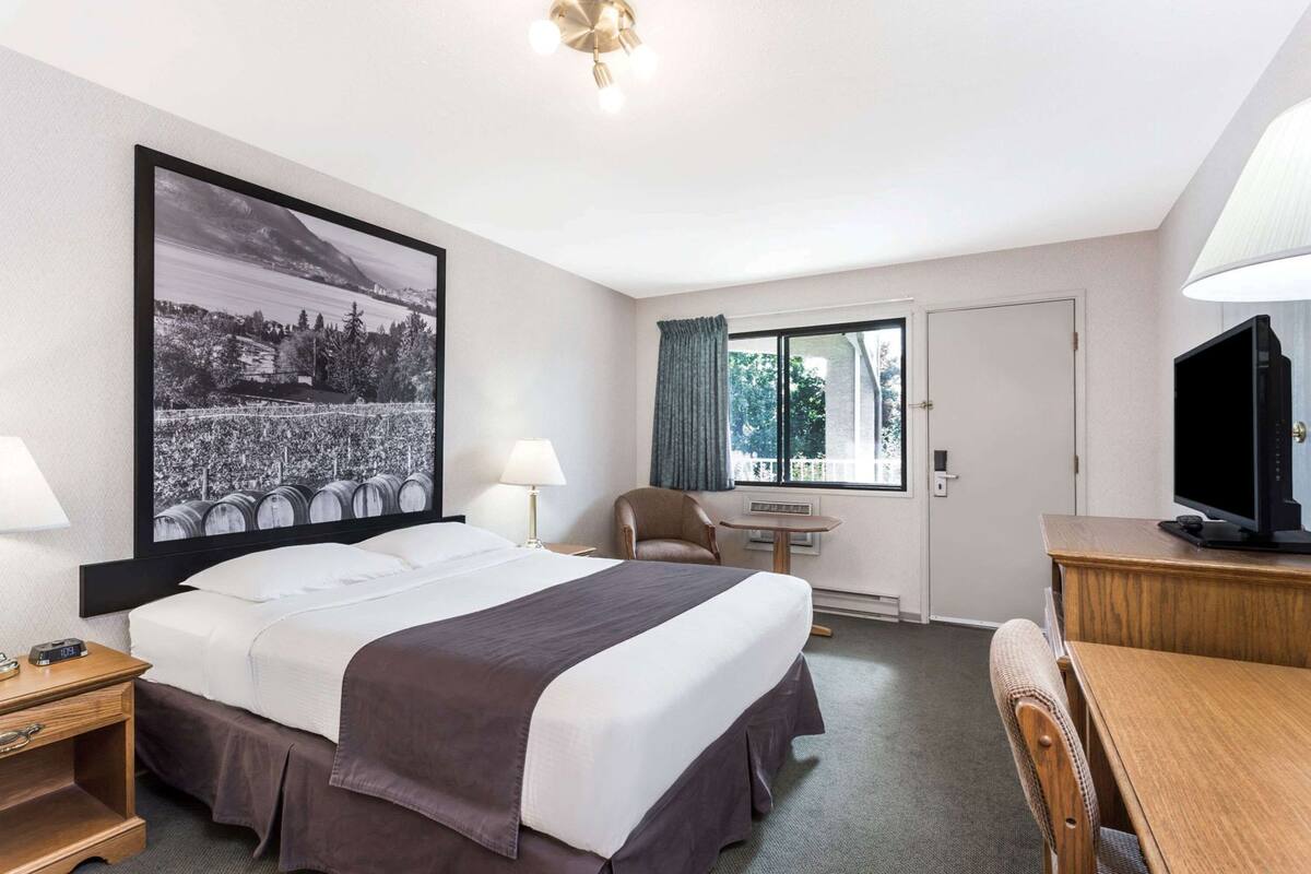 A hotel room with a large bed, a desk, a chair, a television, a window with curtains, and a wall-mounted picture.