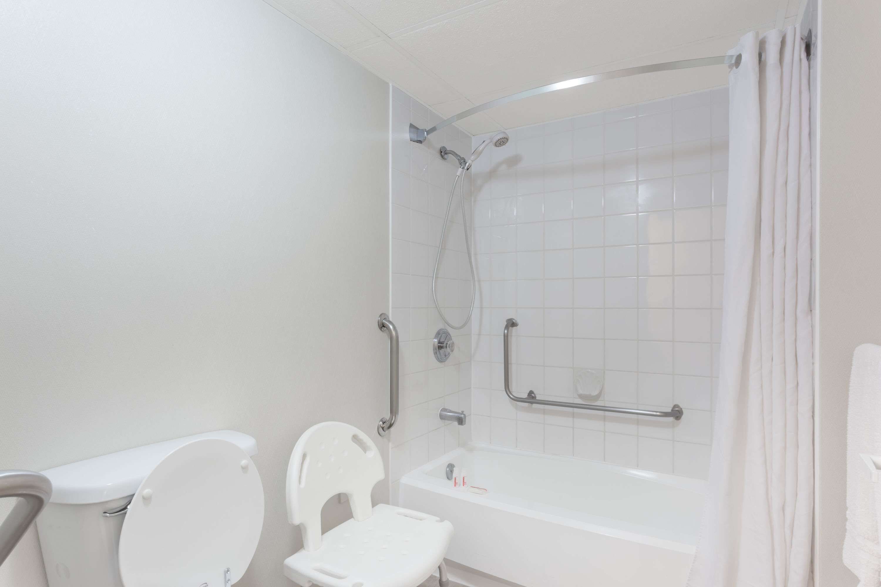 Un baño con inodoro blanco, lavamanos blanco, ducha blanca con barras de apoyo y bañera blanca.