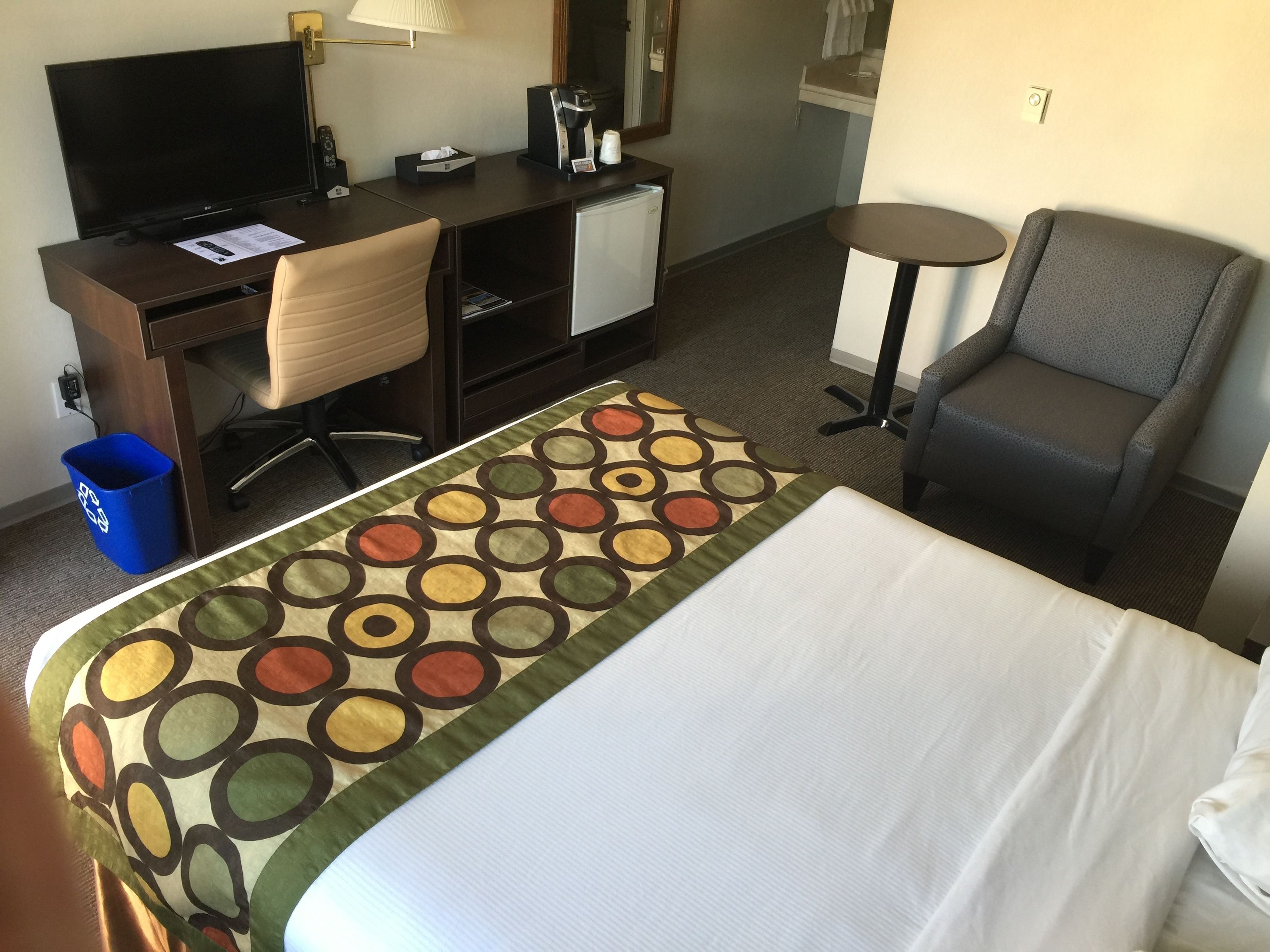Een hotelkamer met een bed, een bureau met een computer, een stoel, een klein tafeltje en een prullenbak.