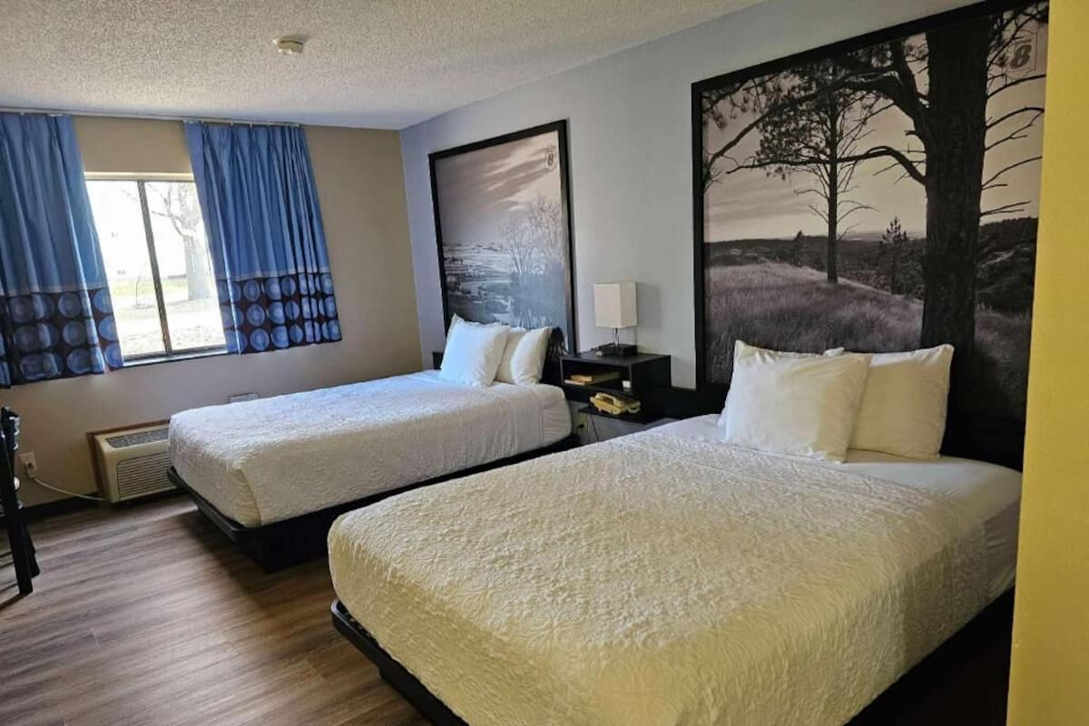 Habitación de hotel con dos camas, un ventanal con cortinas azules, una lámpara de pared y un cuadro grande de paisaje.