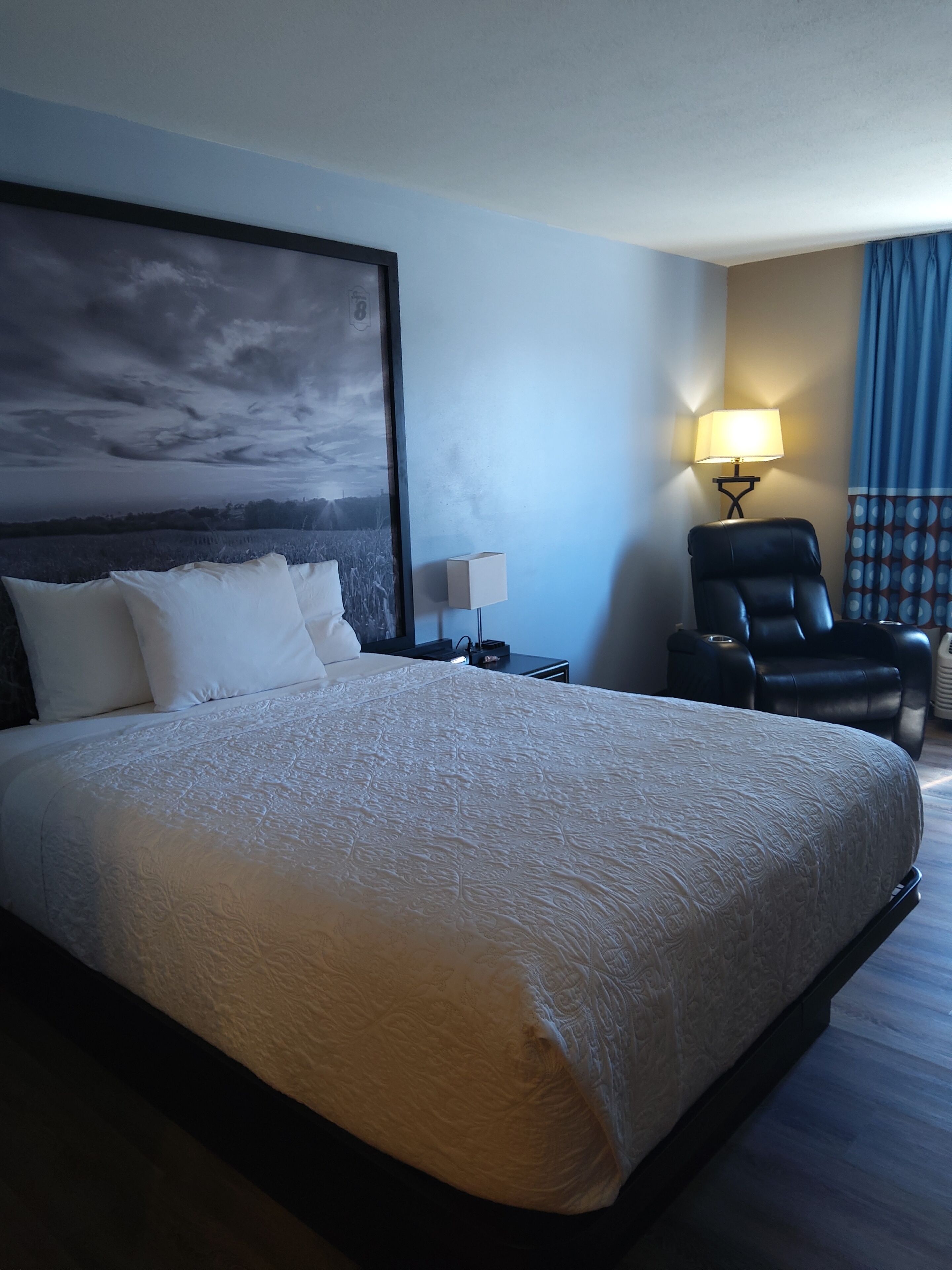 A hotel room with a large bed, a chair, a nightstand with a lamp, and a painting on the wall.