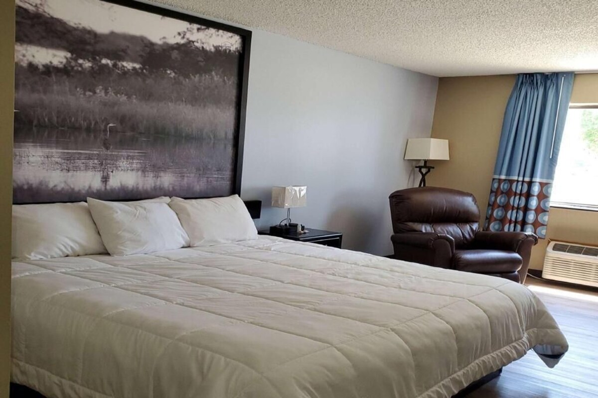 Una cama grande con cubrecama acolchado, una mesita de noche con lámpara, un sillón de cuero y una obra de arte colgada en la pared.