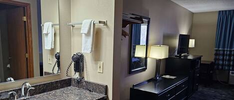 A hotel bathroom with a granite sink, a mirror, a hairdryer, a towel rack, and a desk with a lamp.
