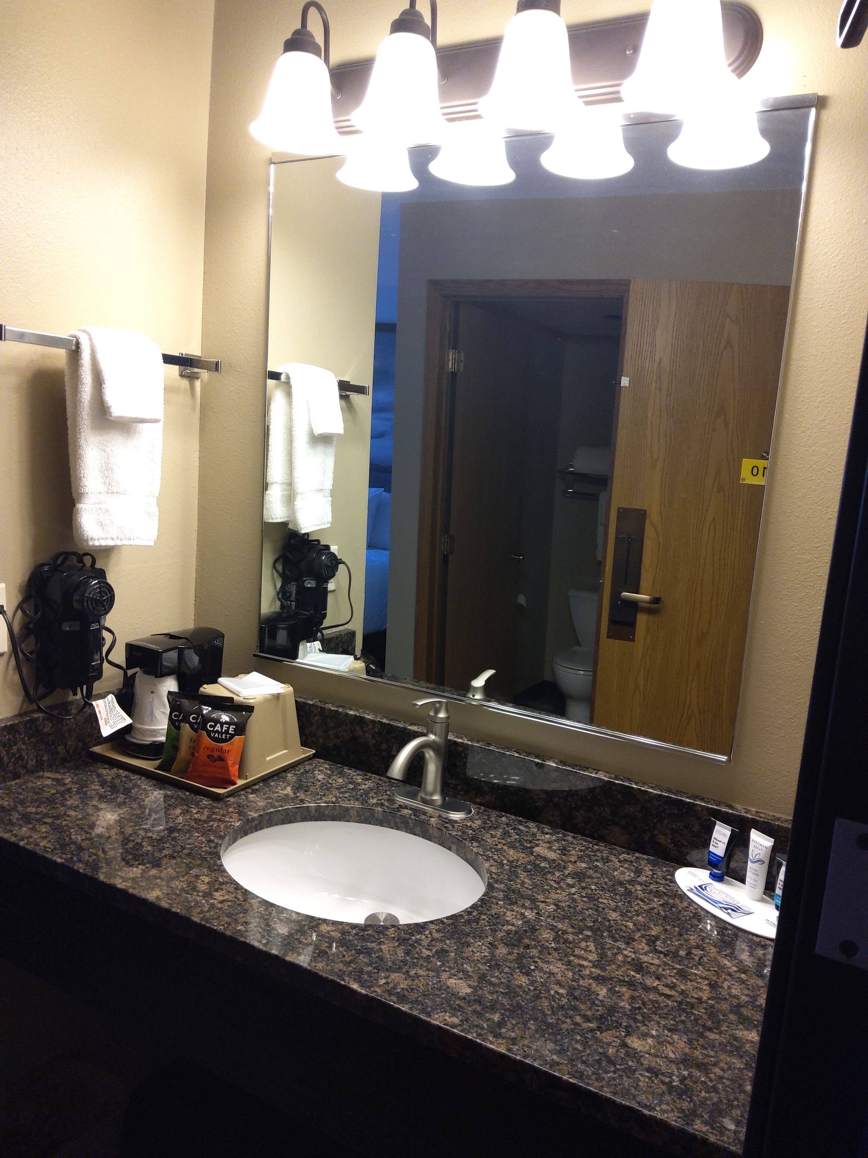 Un baño de hotel con encimera de granito, un lavamanos blanco, una cafetera y un espejo con luces arriba.