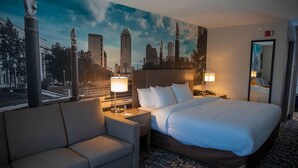 A hotel room with a large bed, two bedside tables with lamps, a sofa, and a cityscape mural on the wall.