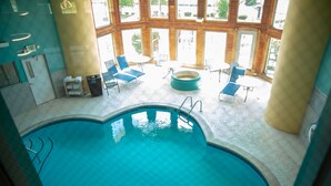 An indoor swimming pool with clear blue water, surrounded by lounge chairs and tables, large windows, and a visible exit sign.