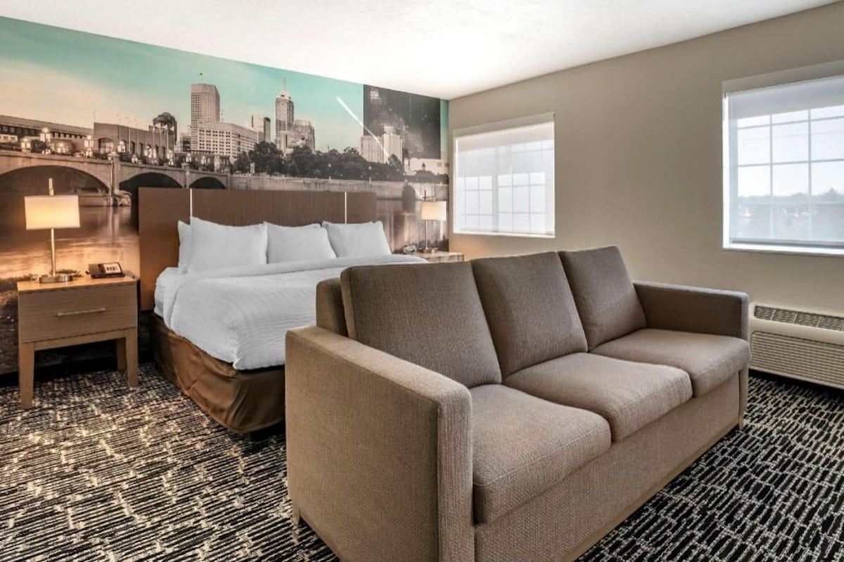 A hotel room with a bed, a sofa, a nightstand, a lamp, a window, and a cityscape mural on the wall.