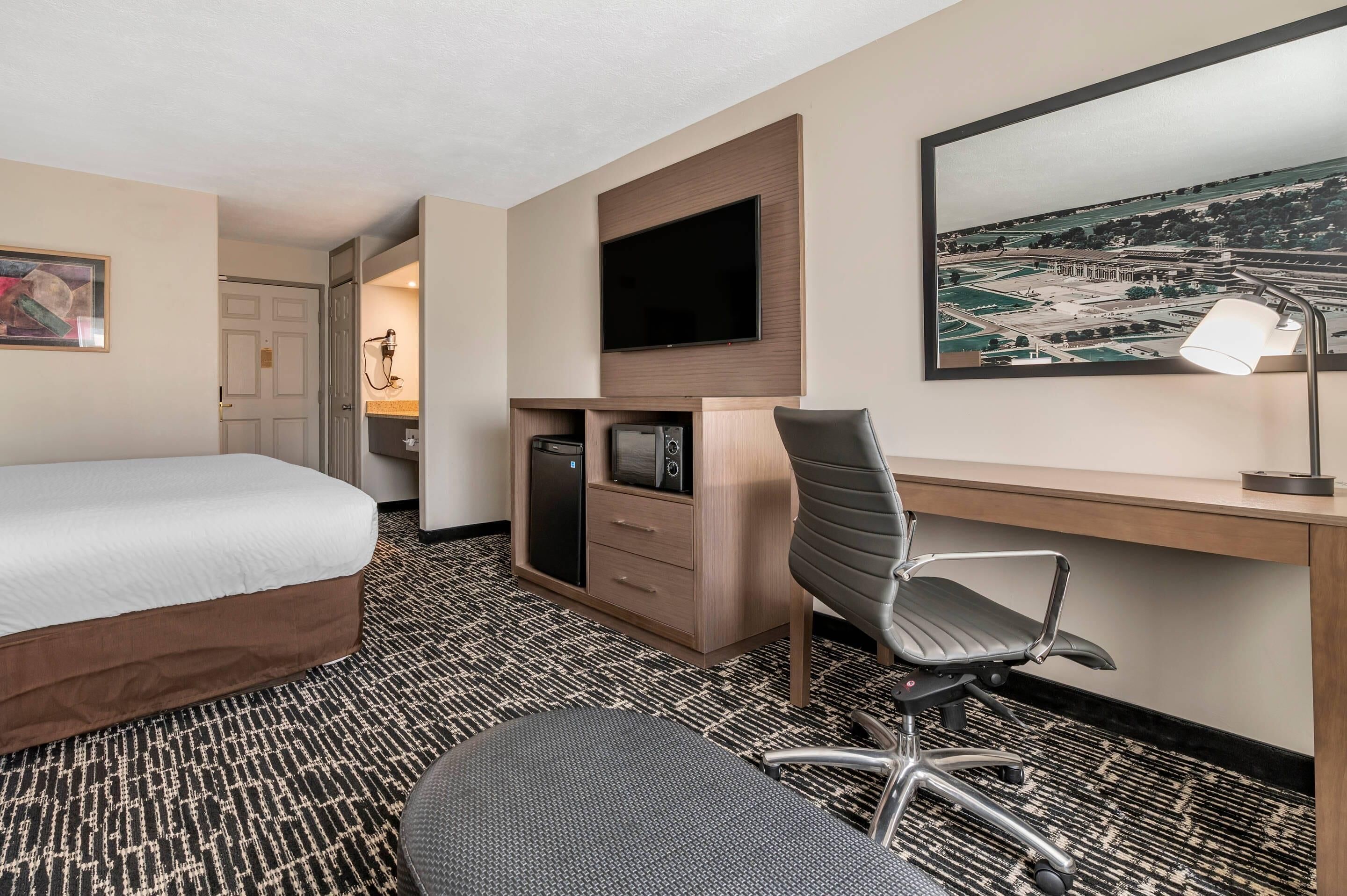 A hotel room with a bed, desk, chair, TV, and artwork on the wall.