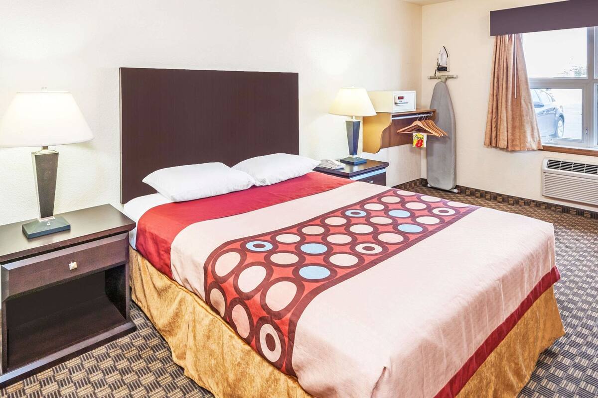 Una habitación de hotel con una cama grande, dos mesitas de noche, una lámpara, una tabla de planchar y un ventanal con cortinas.