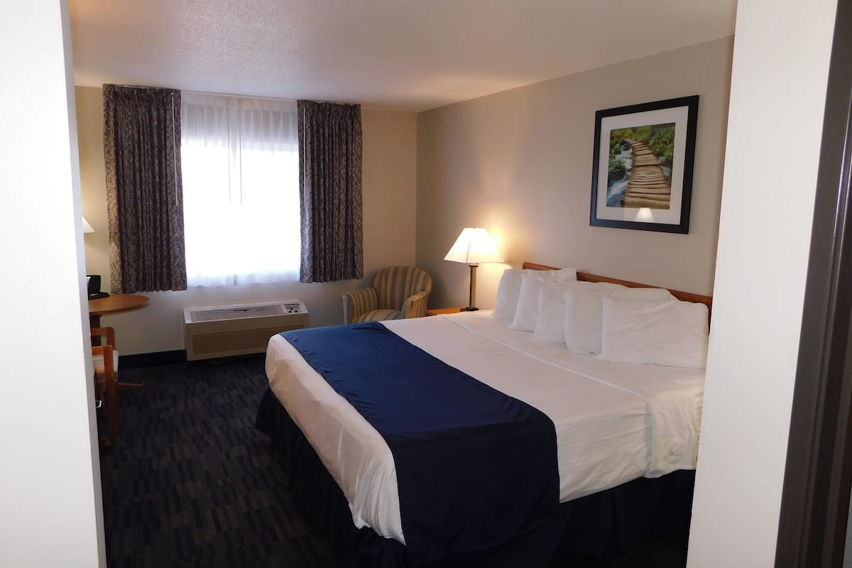 A hotel room with a bed, a desk, a chair, a window with curtains, a wall-mounted lamp, and a framed picture.