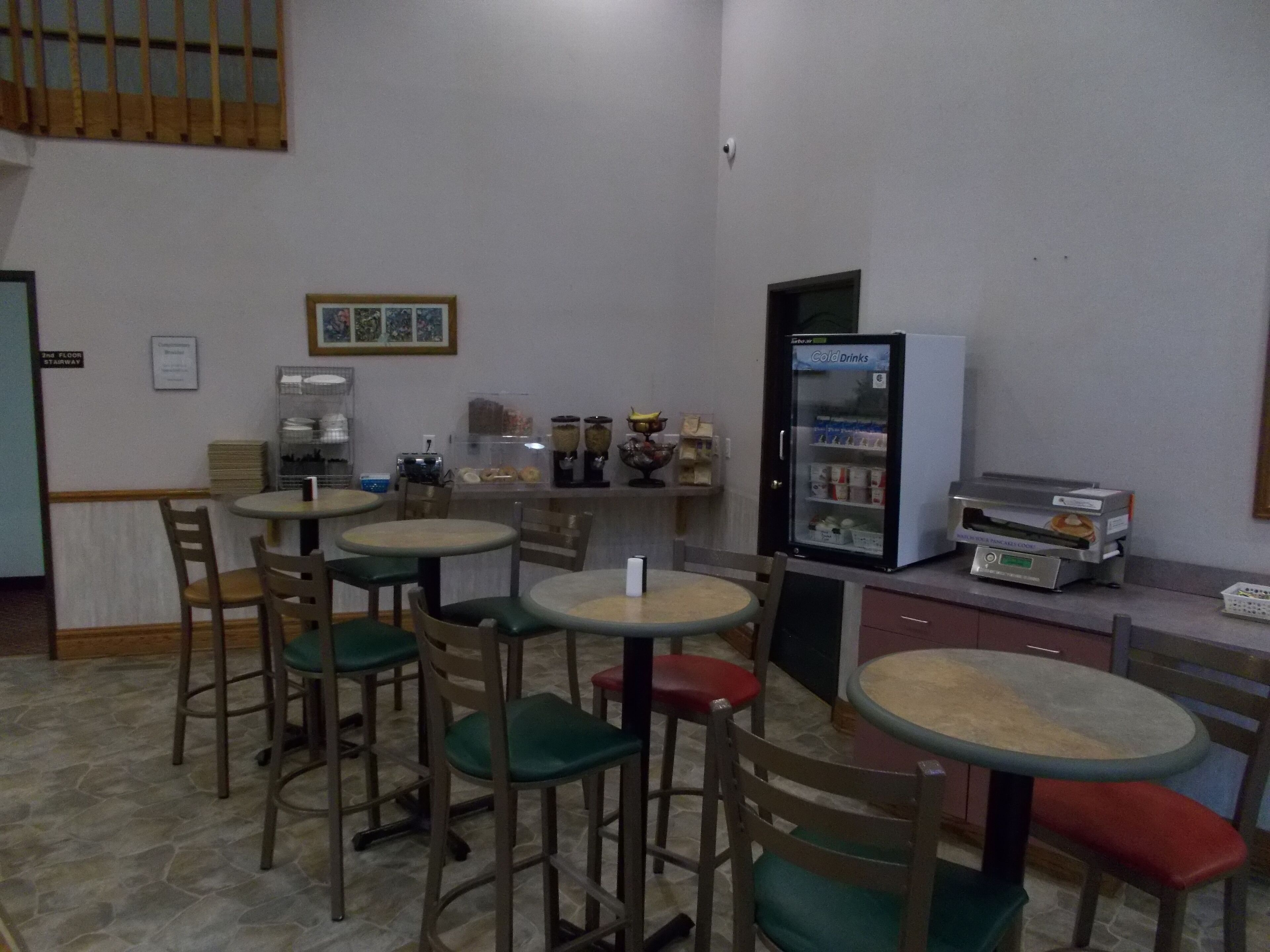 A small cafe with several round tables and chairs, a coffee machine, a refrigerator with beverages, and a sign indicating the location.
