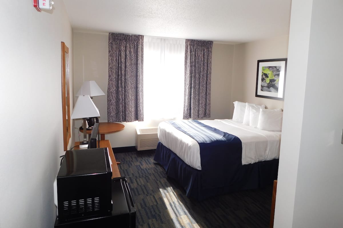 A hotel room with a bed, a desk with a lamp, a chair, a heater, and a fire alarm.