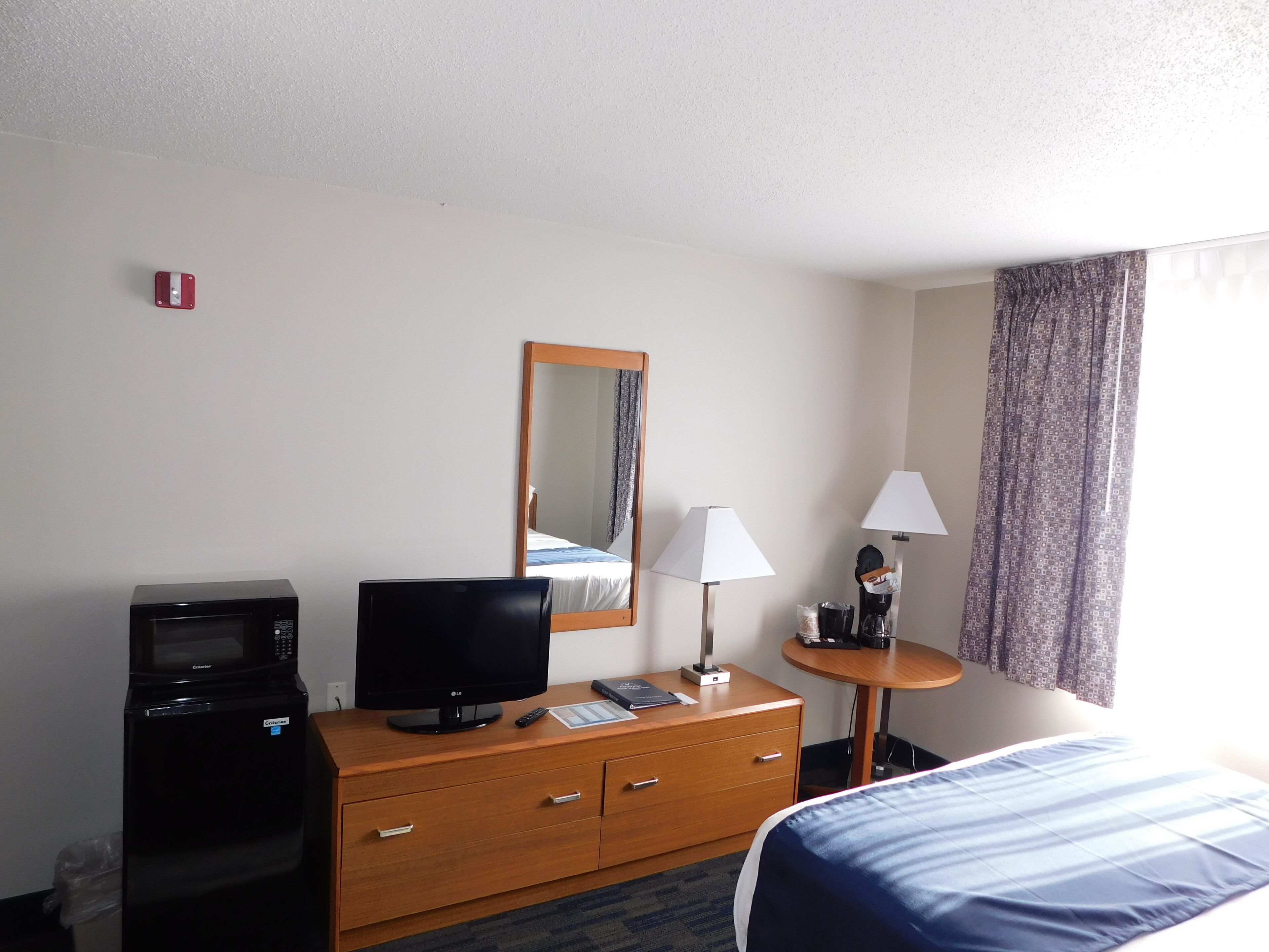 Habitación de hotel con cama, televisor sobre una cómoda de madera, microondas, espejo, lámpara y ventana con cortinas.