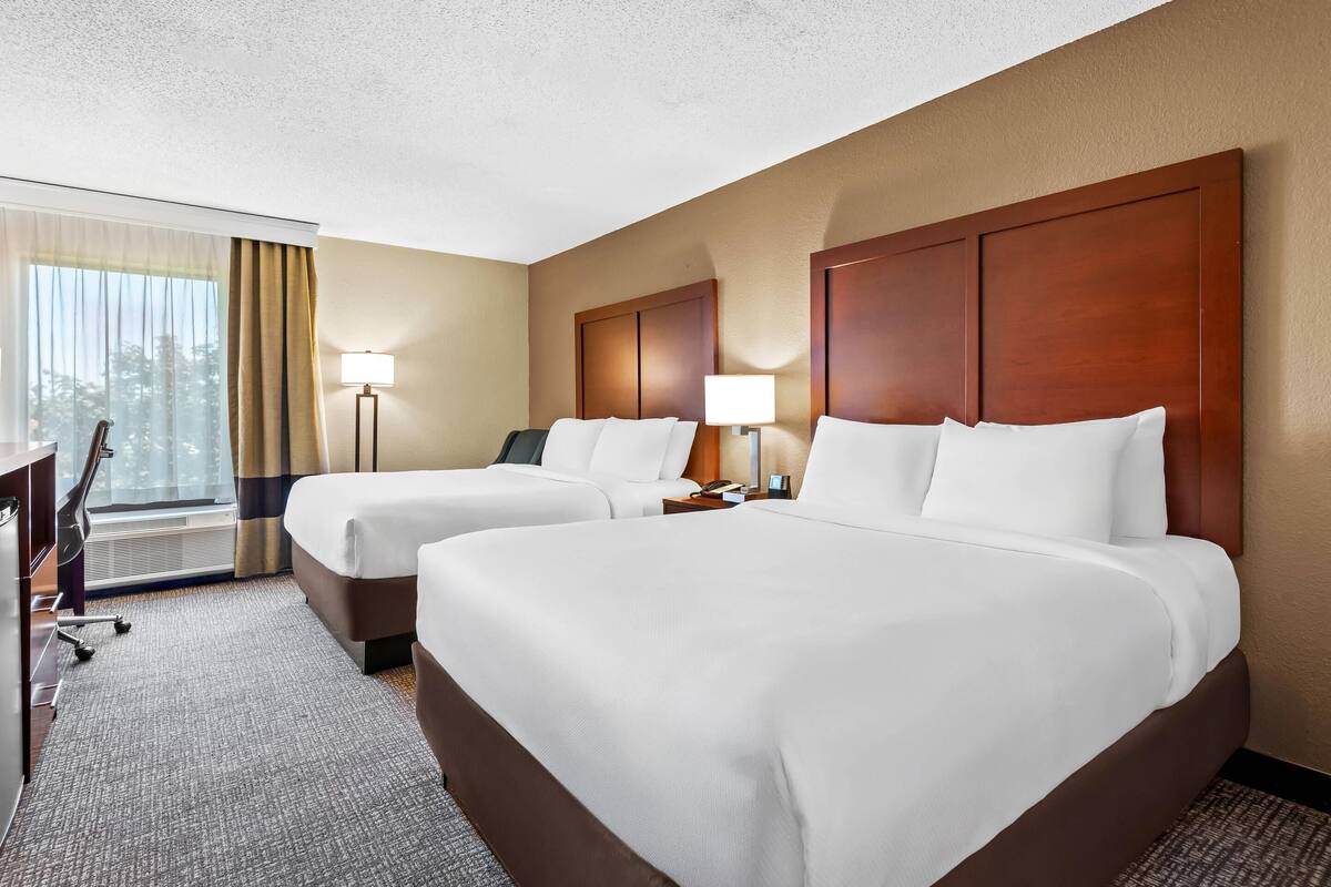 Une chambre d’hôtel avec deux lits, un bureau, une chaise, une lampe et une fenêtre avec des rideaux.