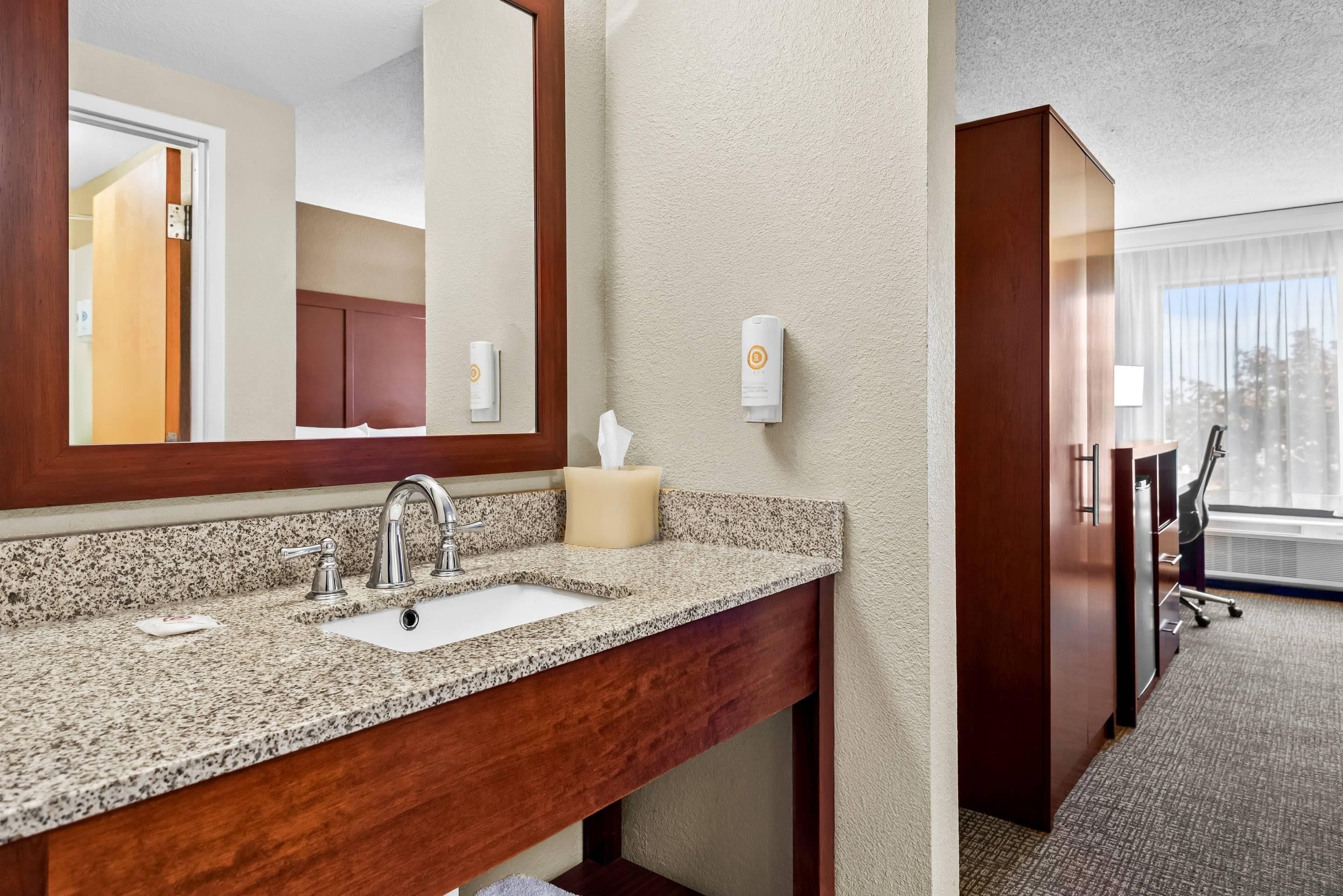 A hotel bathroom with a granite countertop sink, a mirror, and a tissue dispenser.