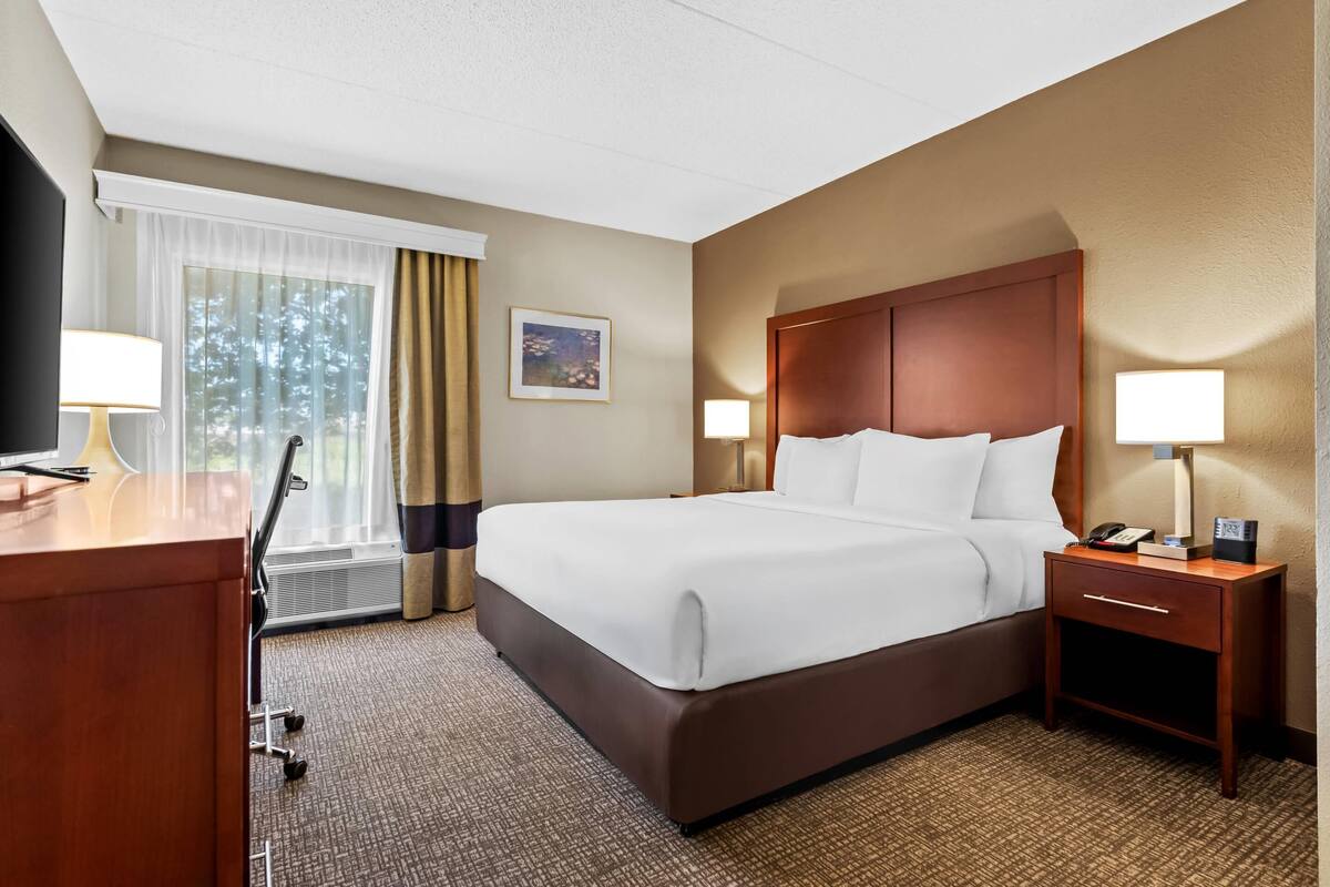Une chambre d’hôtel avec un grand lit, deux tables de chevet avec des lampes, un bureau avec une lampe, une chaise et une fenêtre avec des rideaux.