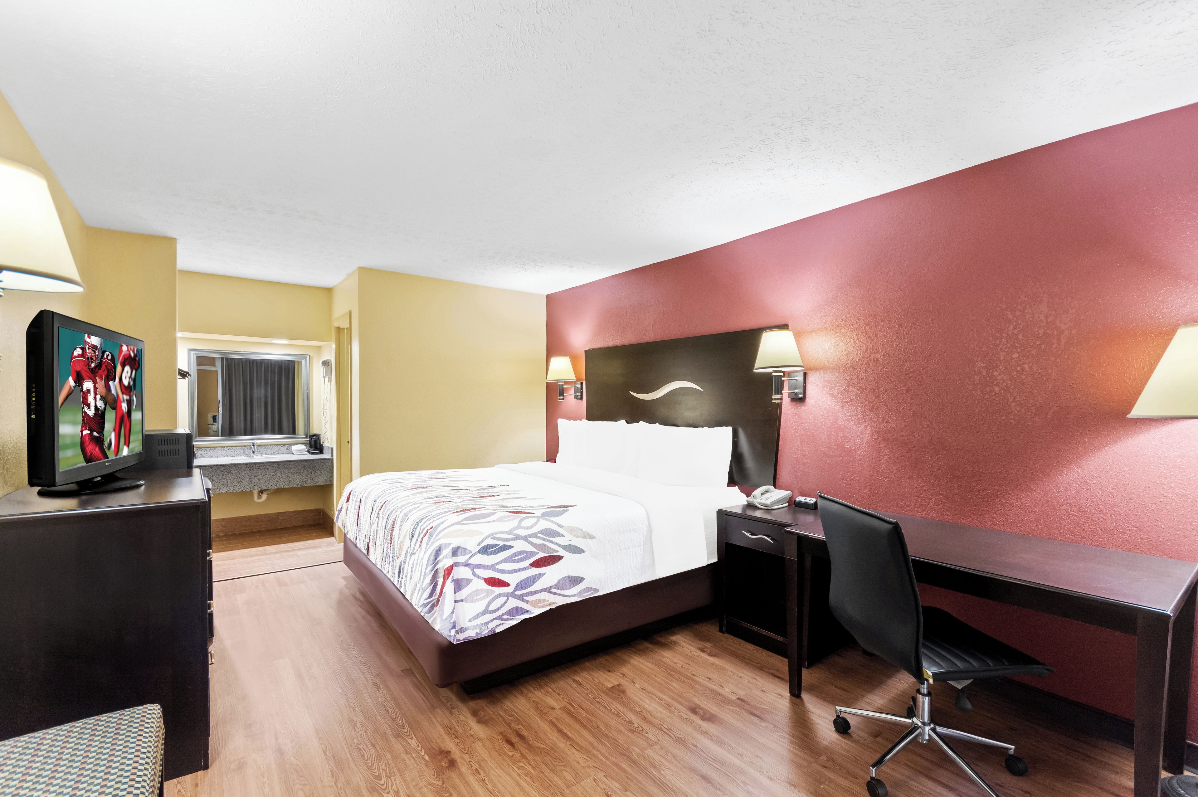 A hotel room with a bed, desk, chair, TV, and lamps.