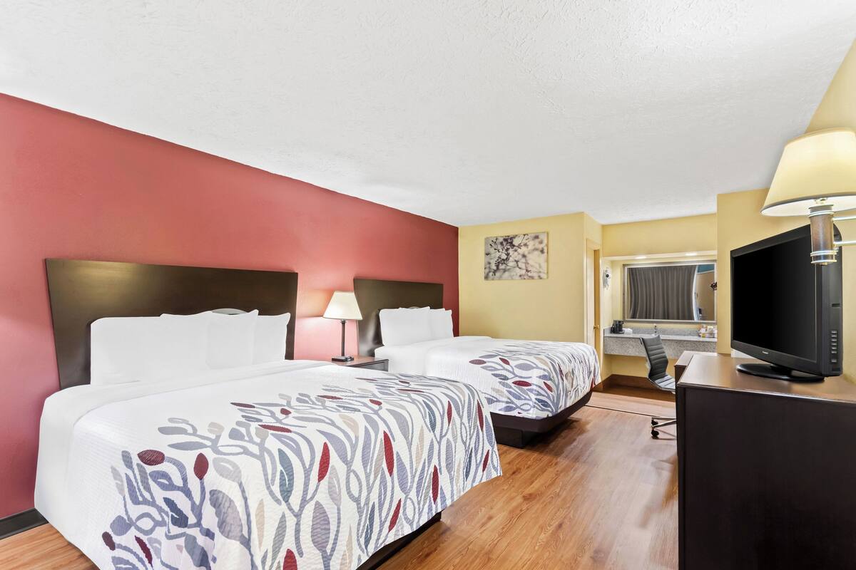 A hotel room with two beds, a desk, a TV, and a window.