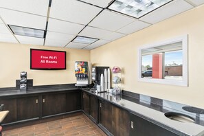 Una hall d'albergo con una televisione a schermo piatto che mostra 'Wi-Fi gratuito in tutte le camere', una macchina del caffè e una finestra con vista sull'esterno.