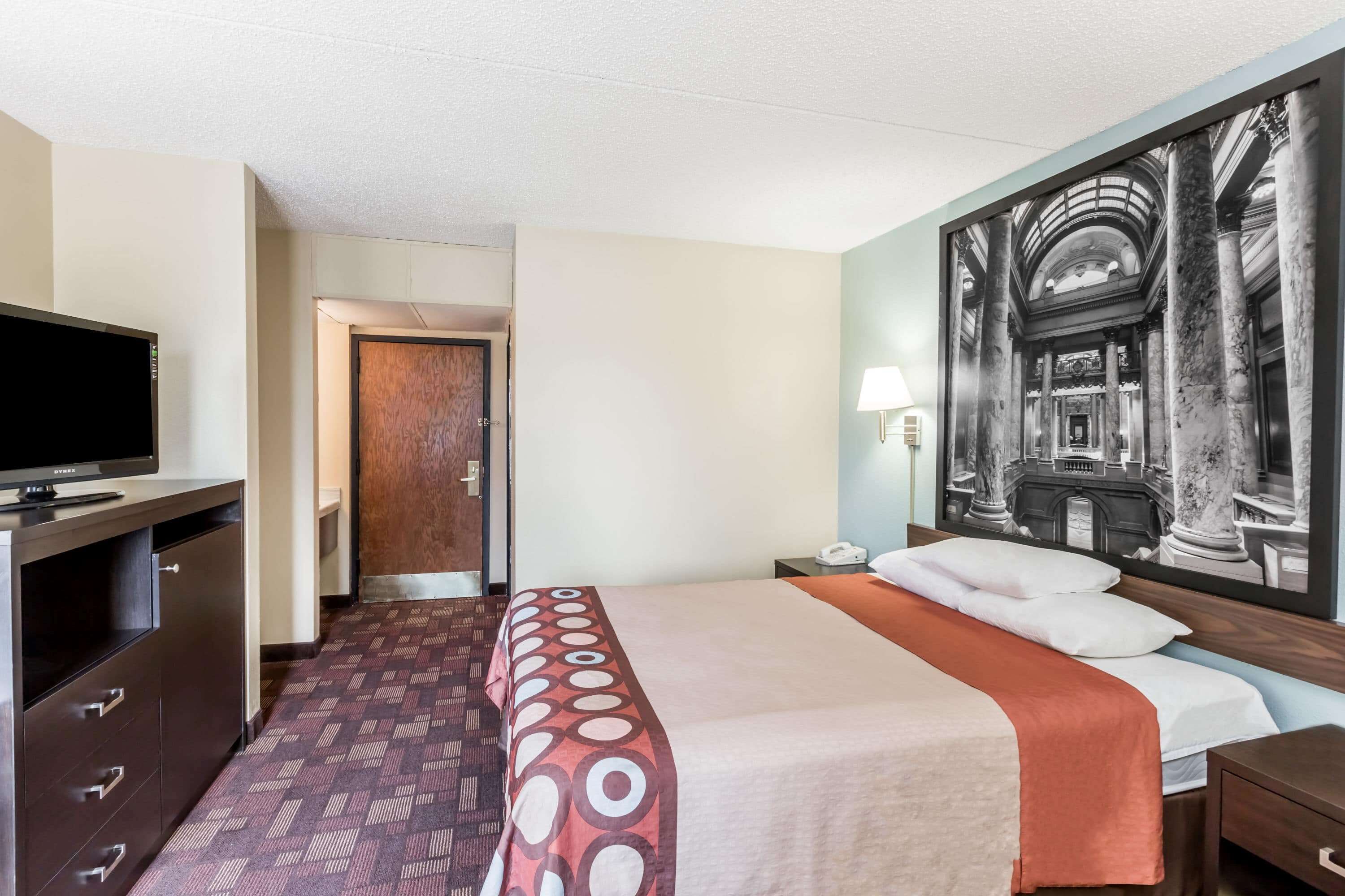 A hotel room with a bed, a desk with a television, a painting on the wall, and a door leading to another room.