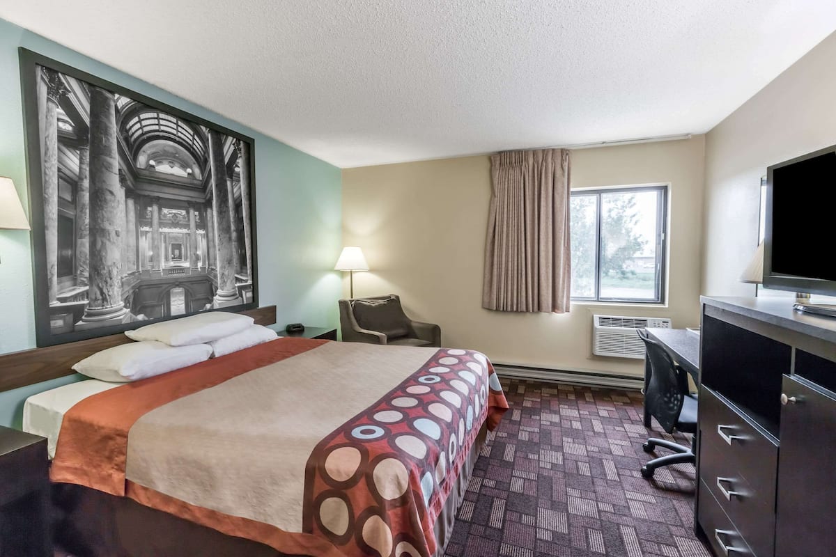 A hotel room with a bed, a desk, a chair, a television, and a large framed picture on the wall.
