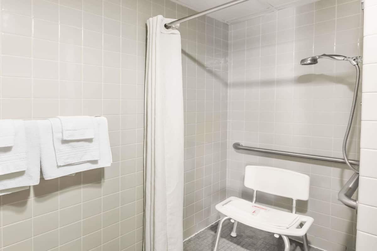 A bathroom with white tiles, a shower with a curtain, a white chair, and a handrail.