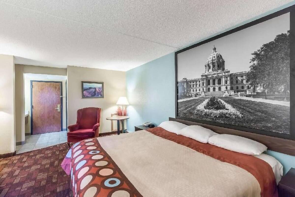 A hotel room with a bed, a red armchair, a small table with a lamp, and a large framed picture of a building.