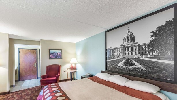 A hotel room with a large bed, a red armchair, a small table with a lamp, and a framed picture of a building.