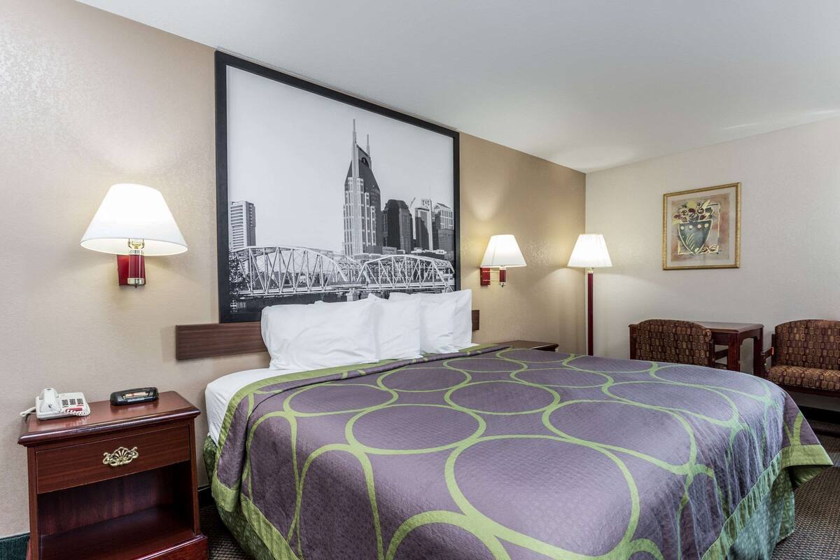 Una habitación de hotel con una cama grande, dos lámparas de noche, una mesita de noche con un teléfono, un cuadro enmarcado de un paisaje urbano y una pequeña mesa con una silla.