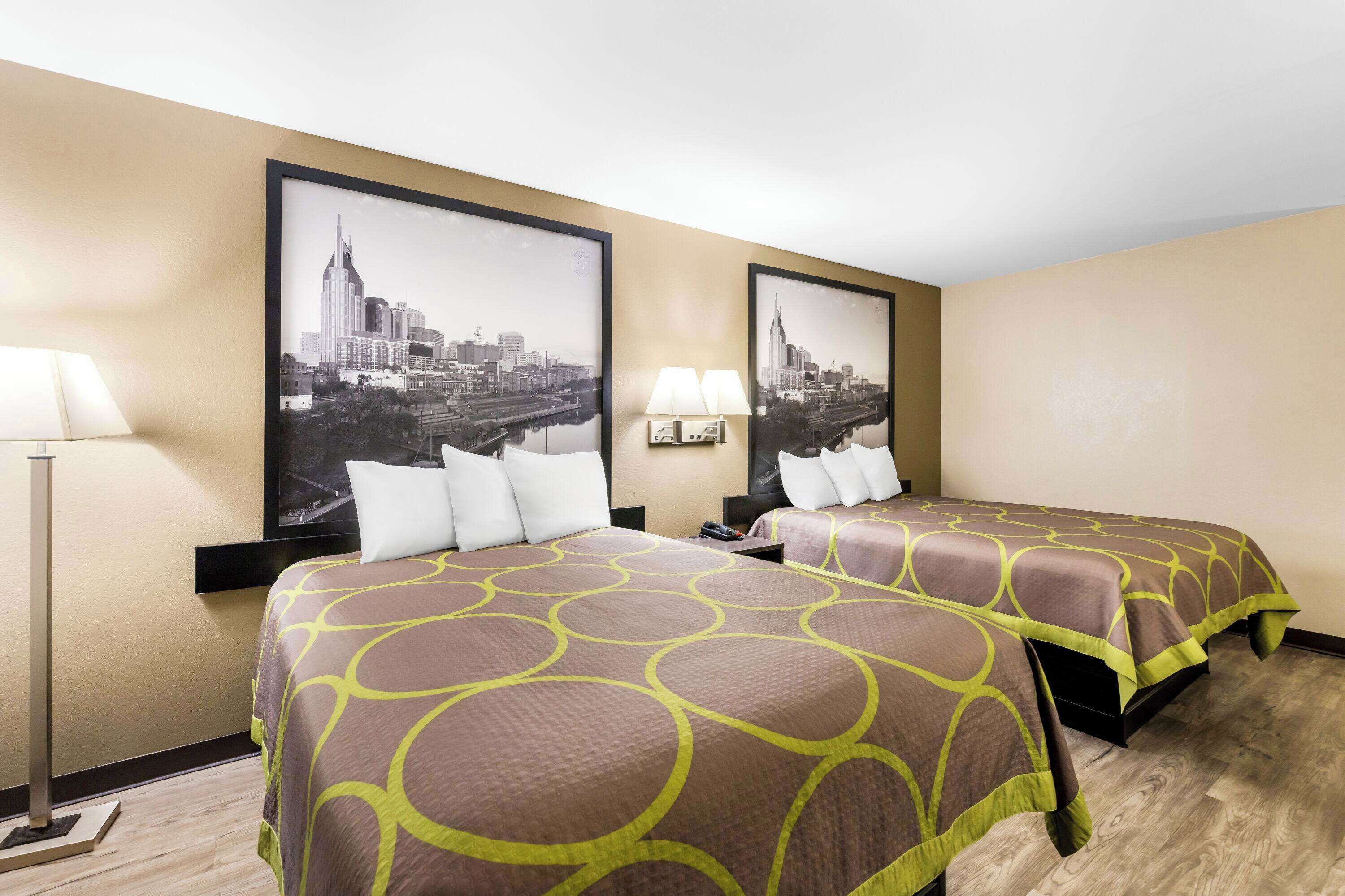 A hotel room with two beds, each with a brown and green patterned bedspread, a cityscape poster on the wall, and a floor lamp.