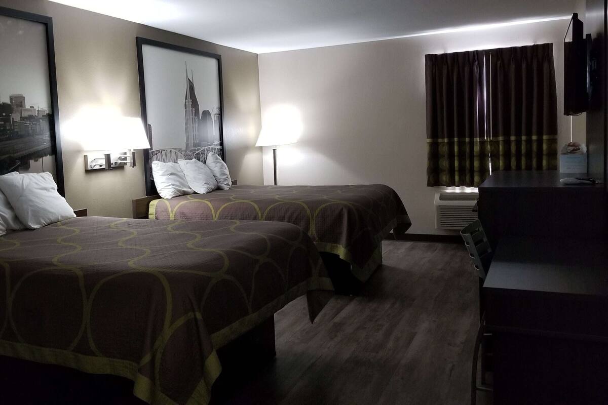 Habitación de hotel con dos camas, un escritorio, un televisor y una fotografía enmarcada de un paisaje urbano.