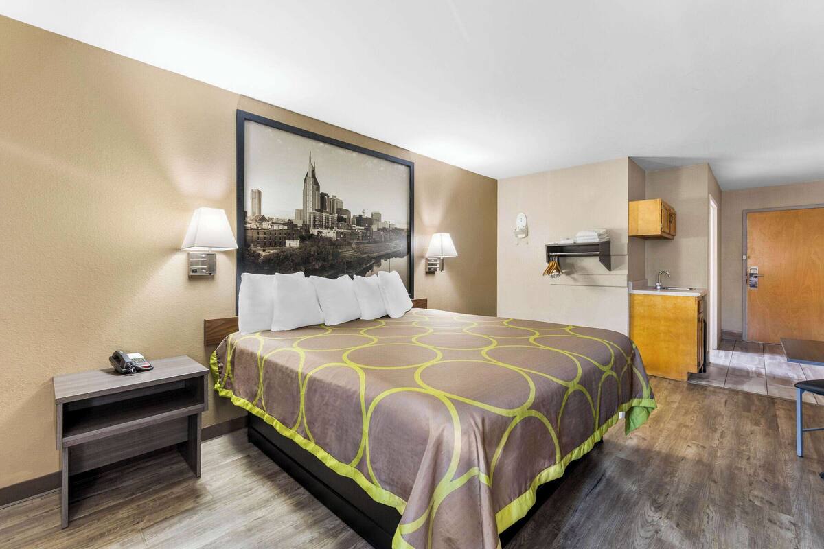 Habitación de hotel con cama, lámparas de noche, mesita de noche, escritorio, silla, kitchenette y puerta.