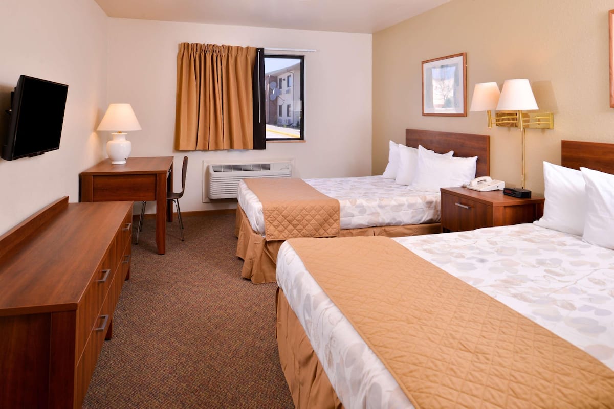 A hotel room with two beds, a desk, a chair, a TV, an air conditioning unit, and a window with curtains.