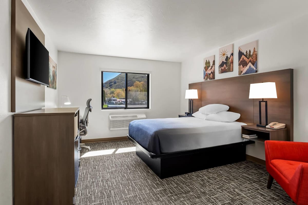 Una habitación de hotel con cama, lámparas de noche, un escritorio con televisor de pantalla plana, una silla y ventana con vista a la montaña.