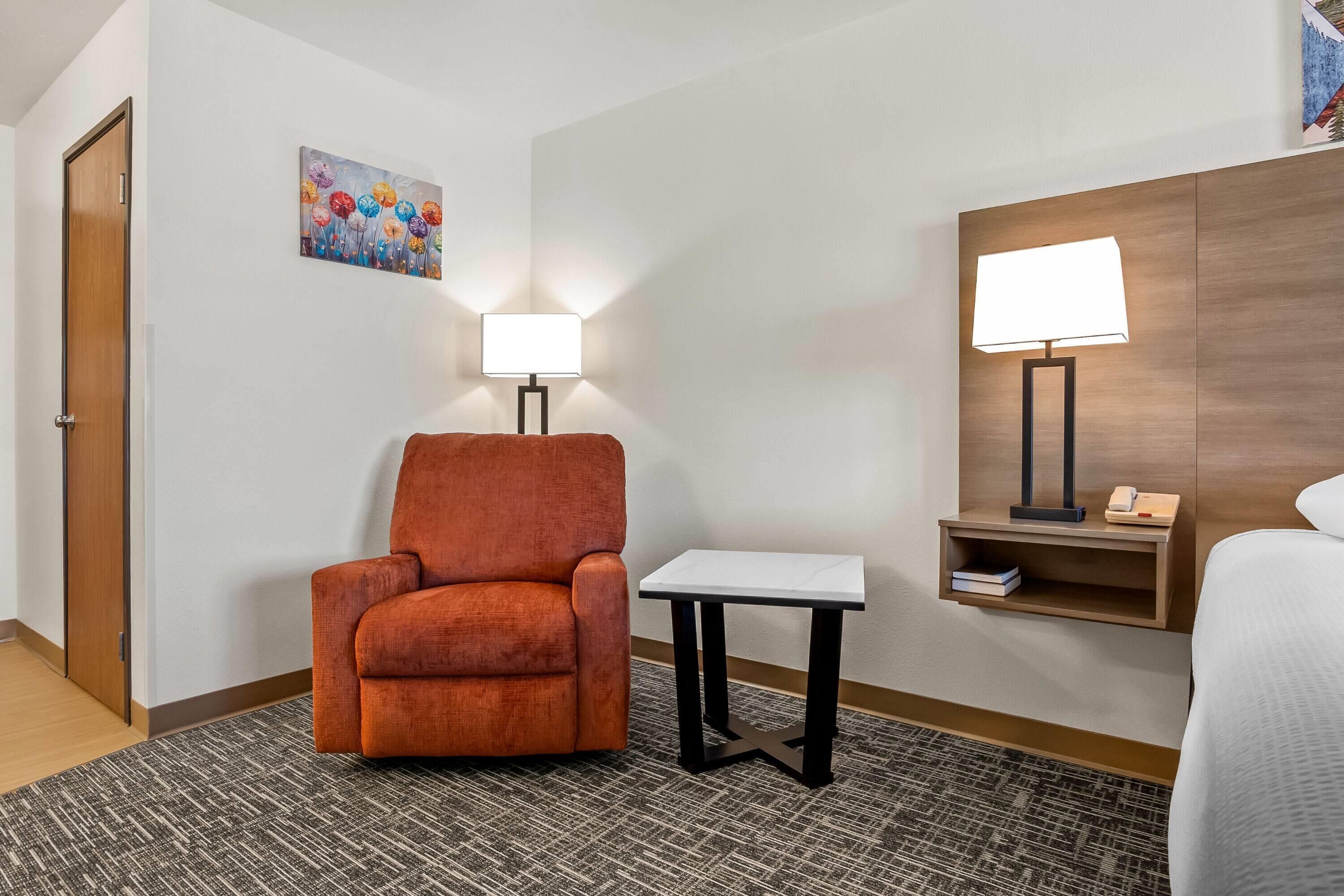 Una habitación de hotel con una única silla naranja, una mesita pequeña, una lámpara, una mesita de noche con lámpara y un cuadro en la pared.
